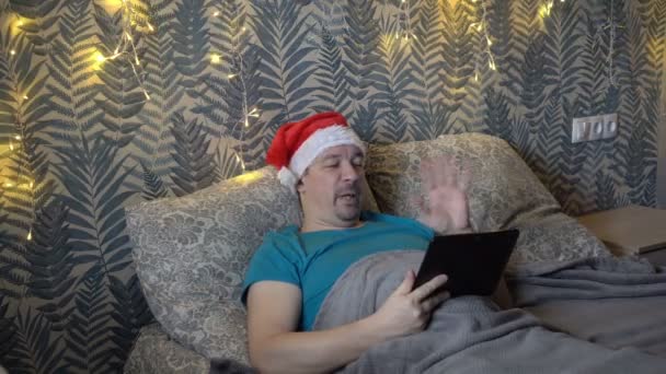 Um homem de meia-idade barbudo se comunica via link de vídeo através de um tablet, deitado em uma cama à noite em um chapéu vermelho de Papai Noel contra um fundo de uma guirlanda piscando amarela. O conceito. Close-up. 4K — Vídeo de Stock