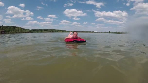 Wesoła dziewczynka unosi się na czerwonym materacu na środku jeziora, daleko od brzegu. Pływa, patrzy w kamerę i uśmiecha się. Zwolnij trochę. Strzelanie z poziomu wody. 4K — Wideo stockowe