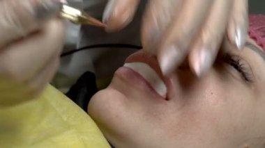 Yakın plan, kız güzellik salonunda kalıcı olarak dudak makyajı yapıyor ve kozmetik uzmanı dudaklarına iğne batırarak boya sürüyor. Bir kozmetolojistin işi. 4K