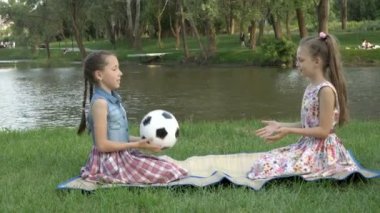 Futbol topuyla oynayan iki mutlu küçük kız. Birbirlerine atıyorlar, gün batımında şehir parkında çimlerin üzerinde nehrin arka planına karşı oturuyorlar. Eğlenceli çocuk tatili. 4K