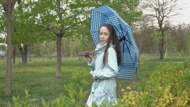 Seorang gadis remaja berambut panjang berpose untuk kamera di sebuah taman kota kosong, dengan payung biru di tangannya terhadap latar belakang tanaman kuning dan hijau pada hari musim semi. Potret. 4K — Stok Video
