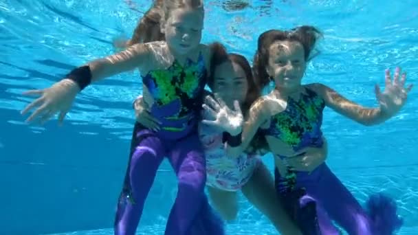 Tres niñas adorables bucean lentamente bajo el agua en una piscina al aire libre en hermosos trajes de baño en un día soleado. Miran a la cámara bajo el agua, sonríen y saludan con las manos. Movimiento lento — Vídeo de stock