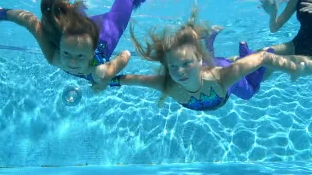 Two happy little girls swim and pose underwater in an outdoor pool in beautiful swimsuits on a sunny day. They look at the camera, smile, and wave. Slow motion. Bottom view. Close-up — Stock Video