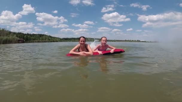 一个小女孩和她的妈妈玩得很开心，在水面上的红色气垫上飘浮着，他们的脚也在飞溅。他们看着相机笑了。慢动作4K — 图库视频影像