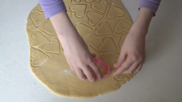 Una niña aprende a hacer galletas. Ella utiliza una plantilla para cortar los espacios en blanco de galletas en forma de corazón de la masa cruda. Primer plano. Sin rostro. Vista superior. 4K — Vídeo de stock