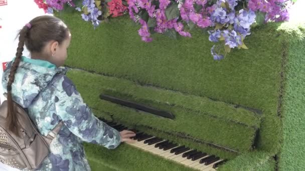 Una joven, una adolescente, en ropa de abrigo, toca el piano, que se encuentra en un lugar público. El piano está decorado con flores y vegetación. Primer plano. Vista trasera. 4K — Vídeo de stock