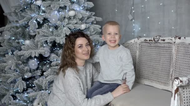 美しい若い母親は笑顔で、装飾されたクリスマスツリーの隣のソファの家に座っている彼女の若い息子を抱きます。彼らは一緒に笑顔と離れて見て。接近中だ。4K — ストック動画