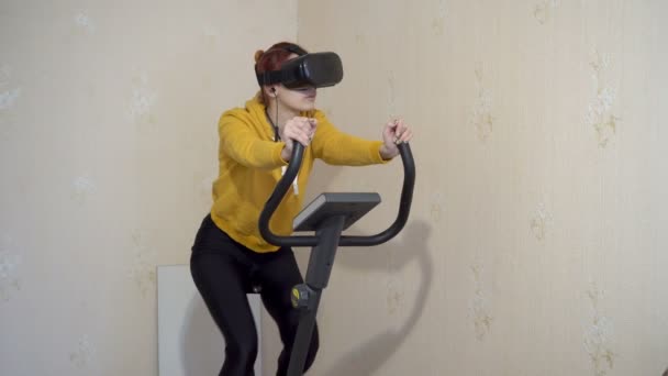 Een jong meisje zit thuis op een hometrainer in een virtual reality bril. Een meisje in gele sportkleding. Thuissporttraining. Een close-up. 4K — Stockvideo