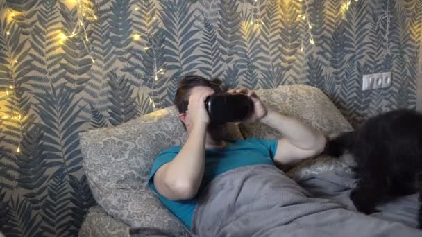 En voksen mand iført virtual reality briller leger med en sød kæledyr, en sort køter hund, mens liggende på en seng derhjemme om aftenen. Nærbillede. 4K – Stock-video