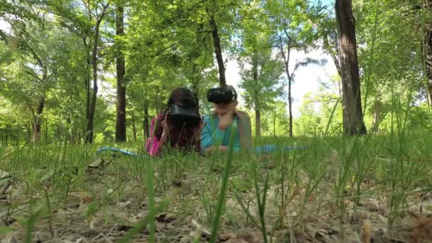 Anya és a kislánya pihennek és játszanak, a parkban fekszenek a füvön virtuális valóság szemüvegben, és körülnéznek. A virtuális játékban játszanak. 4K — Stock videók