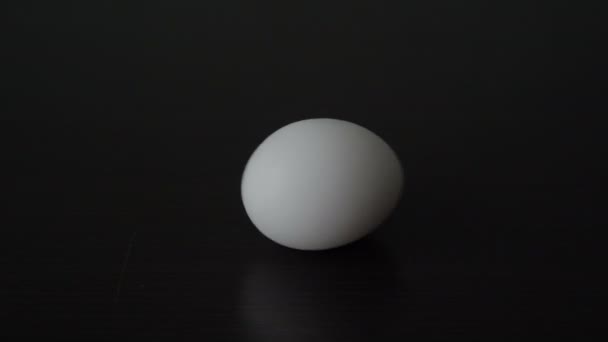 Primer plano, una mano de hombre comienza a rotar un huevo de pollo blanco que está acostado en una mesa negra. Realismo conceptual. Copiar espacio. 4K. — Vídeos de Stock