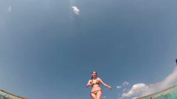 Egy boldog fiatal lány mosolyog, és beugrik a vízbe a mólóról a kamera felett egy napsütéses nyári napon a kék ég felé. Egy fürdőruhás lány átugrik a kamerán. Ugrás, alsó nézet. 4K. — Stock videók