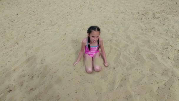 Una bambina abbronzata e felice si siede sulla sabbia sulla spiaggia, sorride, guarda la telecamera, e spruzza sabbia gialla sui piedi. Il concetto di una vacanza felice e spensierata. Vista dall'alto. Ricevuto. 4K. — Video Stock