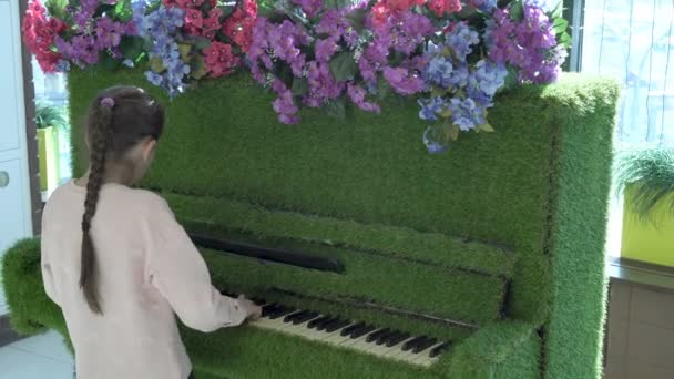 Una bambina carina si diverte e suona a casa su un insolito pianoforte verde, decorato con fiori ed erba. Primo piano. Vista posteriore. 4K. — Video Stock