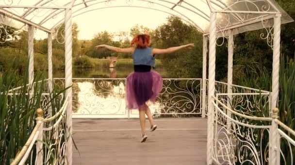 Una bella ragazza si sta divertendo, saltando, ridendo e agitando le braccia su un molo di legno vicino a uno stagno in un parco cittadino al tramonto in una giornata estiva. Ragazza felice con i capelli rossi in una gonna viola. 4K. — Video Stock