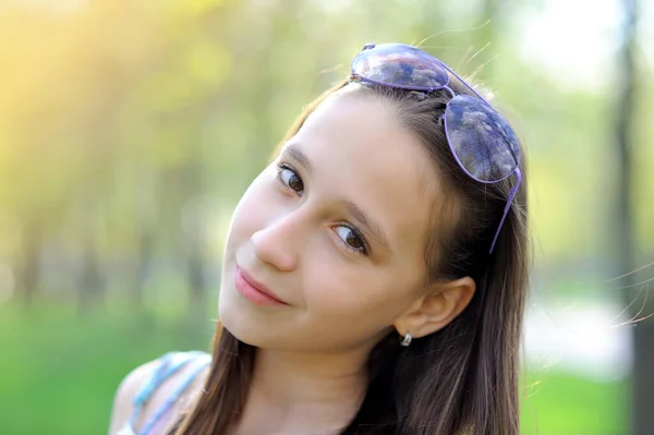 Portrait of a charming young girl in a park at sunset, looking at the camera and smiling sweetly with sunglasses on her head. Close-up. Horizontal orientation of the image. — Stock Photo, Image