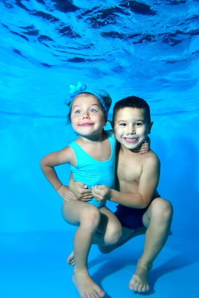 Los niños, un niño y una niña, sonríen y posan bajo el agua en la piscina sobre un fondo azul. Retrato. Primer plano. Orientación vertical. — Foto de Stock