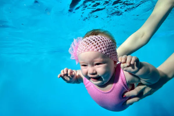 Un bebé llorando en los brazos de su madre bajo el agua en la piscina de los niños. Mamá la pone bajo el agua. Una niña en traje de baño rosa con un lazo en la cabeza. Retrato. Concepto. — Foto de Stock