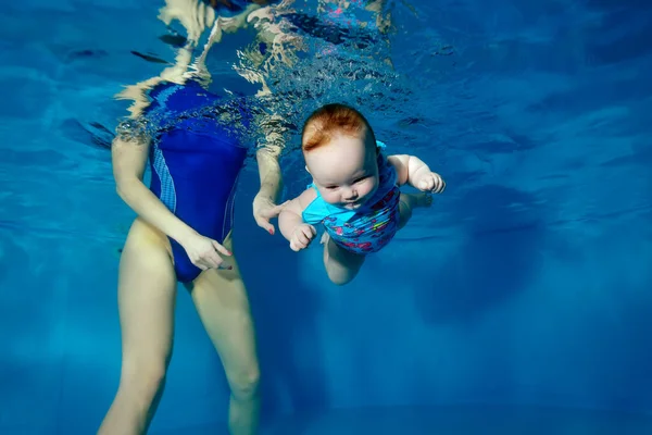 Un bebé bucea y nada bajo el agua en una piscina infantil con los ojos abiertos. Mamá-entrenador la asegura en el agua. Estilo de vida saludable. Natación recreativa para niños. Concepto. Orientación horizontal. — Foto de Stock