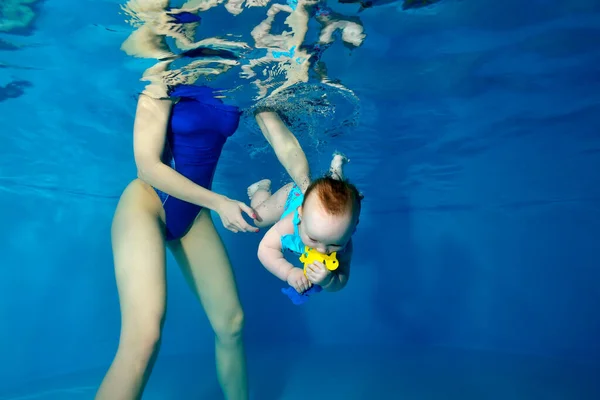 Una niña se sumerge bajo el agua en una piscina para niños con un juguete amarillo en las manos. Una instructora la apoya en el agua. Entrenamiento de natación infantil. Concepto. Orientación horizontal. — Foto de Stock