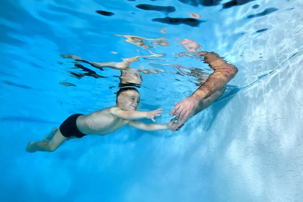 Un niño sonriente nada bajo el agua en la piscina y alcanza las manos de sus papás, que bajó bajo el agua para atrapar al niño. Retrato. El concepto. Orientación horizontal. — Foto de Stock