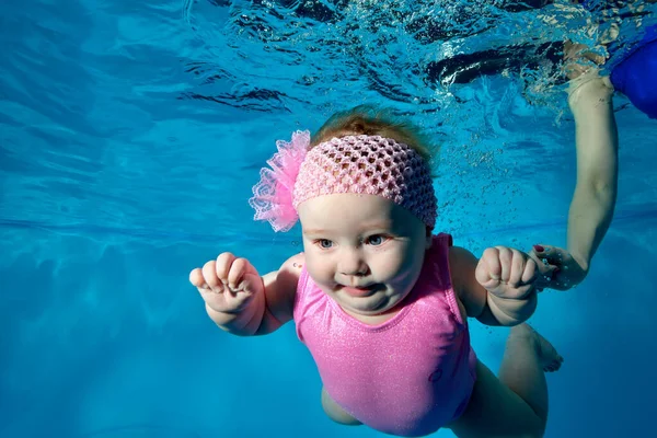 Retrato de un encantador bebé amamantado que nada bajo el agua en una piscina infantil en un traje de baño rosa y con un lazo en la cabeza. Mamá-entrenador la asegura en el agua. Estilo de vida saludable. Concepto. — Foto de Stock