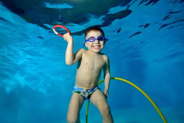 Divertido niño nada y posa bajo el agua en la piscina con un juguete en la mano. Él mira a la cámara y sonríe ampliamente. Retrato. Vista horizontal. — Foto de Stock