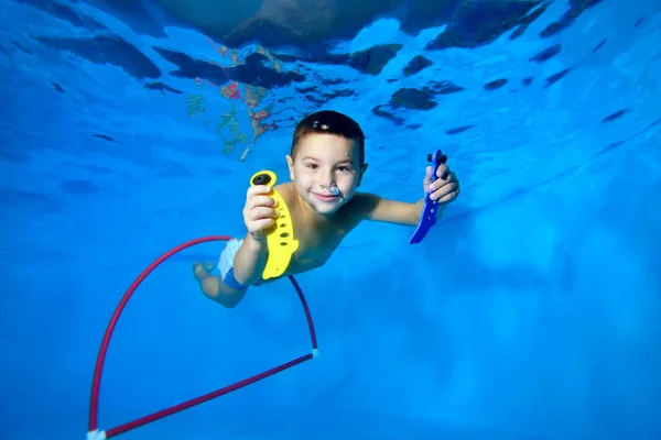 Un niño lindo y sonriente nada bajo el agua en una piscina para niños con juguetes en las manos y mira a la cámara. Retrato. Concepto. Orientación horizontal de la foto. — Foto de Stock