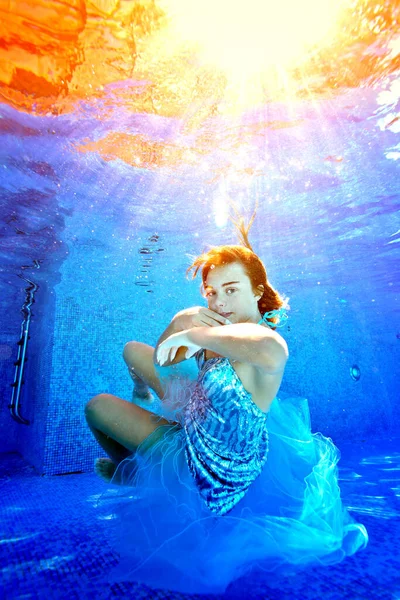 一个玩世不恭的小女孩的画像，她穿着闪亮的蓝色连衣裙，在阳光灿烂的阳光下，在游泳池里的水下对着相机游来游去和摆姿势。概念。纵向方向. — 图库照片