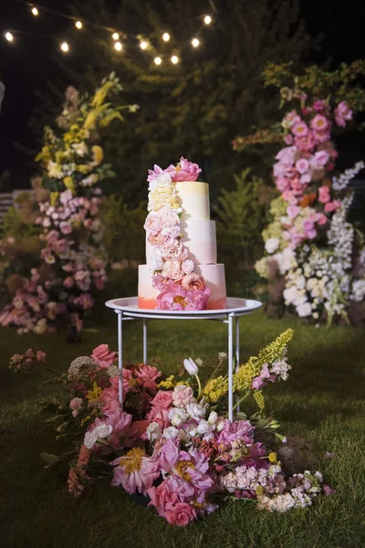 Amazing colorful wedding cake decorated with various fresh flowers standing on white round table outdoors. Dark atmosphere around. Traditional part of wedding celebration.