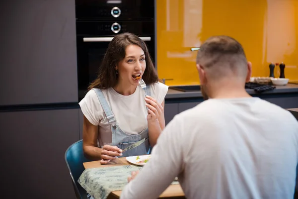 Happy wife and husband sitting together at kitchen table and eating tasty meal. Young couple having breakfast at home. Family lifestyle moments.