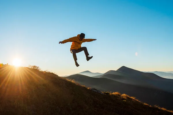 Cheerful young man in casual outfit jumping on peak of mountain. Active hiker enjoying journey on nature. Amazing summer sunrise on background.