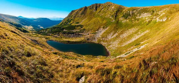 Panoramic view of small lake among high powerful mountains. Blue sky and green nature. Natural landscape.