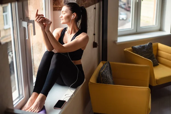 Relaxed woman in black sport clothes enjoying music while sitting near window. Fitness brunette using modern smartphone during break between exercises.