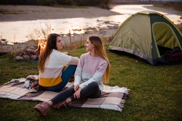 Two women near tent and campfire. Friends are sitting on the grass on a blanket and talking. Enjoy the scenery of mountains and rivers. Hiking, vacation and friendship concept.