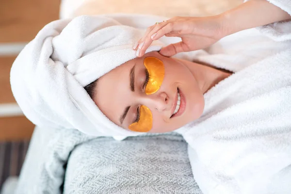 Smiling european girl with under eye patch on face. Young beautiful woman wear bathrobe and wrapped bath towel on head and lying on bed. Close up view. Concept of face skin care