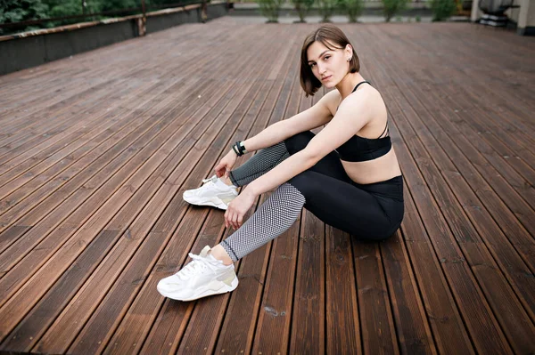 Sexy european sportswoman sitting on wooden terrace or balcony. Young beautiful girl wear sportswear and sneakers and looking to camera. Concept of sport at outdoors. Daytime