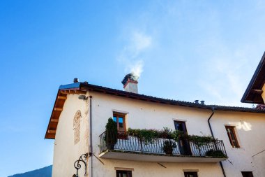 Kır evinde bitkilerle dolu bir balkon, bacadan yükselen duman, açık mavi gökyüzünün altında, huzurlu kırsal mimariyi ve davetkar atmosferi ön plana çıkarıyor.