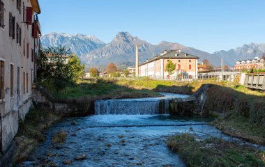 Tranquil river flows through green landscape, surrounded by mountains and buildings, creating a peaceful atmosphere and highlighting the beauty of nature