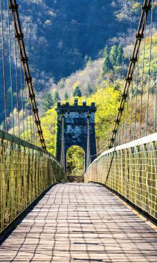 Asma bir köprünün ahşap geçidi canlı yeşillikler ve dağlarla çevrili taş bir kuleye çıkıyor ve güzel bir açık hava manzarası yaratıyor.