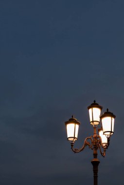 Antika sokak lambası akşam gökyüzünde sıcak bir ışık yayıyor, dekoratif tasarımını vurguluyor ve fotokopi uzayıyla sakin bir atmosfer yaratıyor.