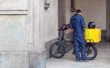Padova, İtalya - 14 Nisan 2025: Mavi kıyafetli teslimat işçisi sarı Glovo kutulu elektrikli bisikletin yanında duruyor.