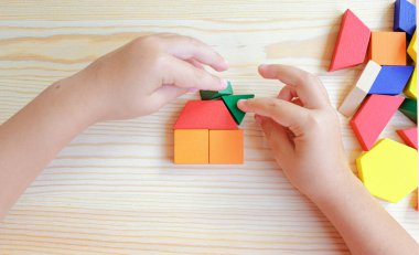 A child's hand constructs a house using multicolored wooden blocks and construction figures. A close-up of multicolored wooden blocks against a wooden table.