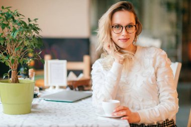 Beyaz süveterli, gözlüklü, kafede oturan ve kameraya pozitif yüz ifadesiyle poz veren güzel sarışın kadın..