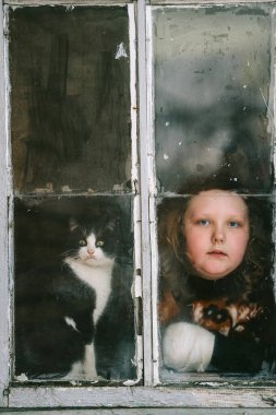 Sıkıcı yüzlü ve sevimli kedisi Coronavirus covid 19 pandemia karantinası sırasında pencereden dışarı bakan küçük bir kızın portresi. Evde kal.. 