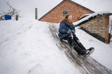 Neşeli kız, naylon poşeti kızak olarak kullanıyor ve bodrumdan iniyor. Kış günü, tuğla köy evinin önünde buz gibi bir yürüyüş yapıyor..