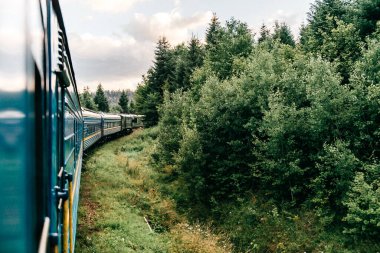 Yazın trenle gezmekten pencere dışındaki manzara. Tepeler, dağlar ve ormanlar. Tatil ve seyahat konsepti. Tren vagonlu lokomotif tren yolu boyunca ilerliyor.