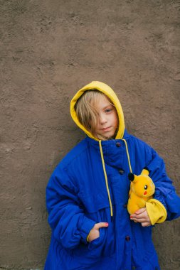 Minsk, Belarus - 8 Aralık 2019: Sıcak mavi ceketli güzel beyaz kız ve küçük oyuncak pikachu 'lu sarı kapüşonlu kız gri bir duvara karşı duruyor
