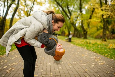 Kalın elbiseli genç anne, sonbahar parkında beyaz oğluyla oynuyor..