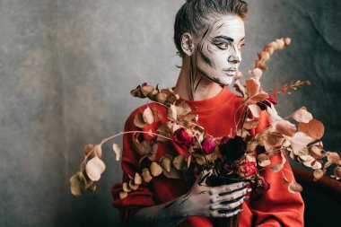 Yaratıcı çizimleri olan güzel genç bir kadın elinde vazoyla arka plandaki grunge duvarının üzerinde kurumuş çiçeklerle (karikatür iskeleti).